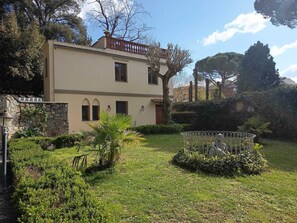 Un edificio de dos plantas con un muro de piedra, un balcón y un jardín bien cuidado con una cerca metálica y diversas plantas.
