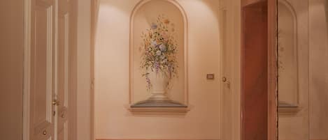 A hallway with wooden flooring, white walls, and a floral arrangement on a pedestal.