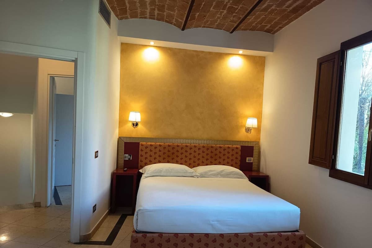 A hotel room with a bed, two bedside lamps, a window, and a door.