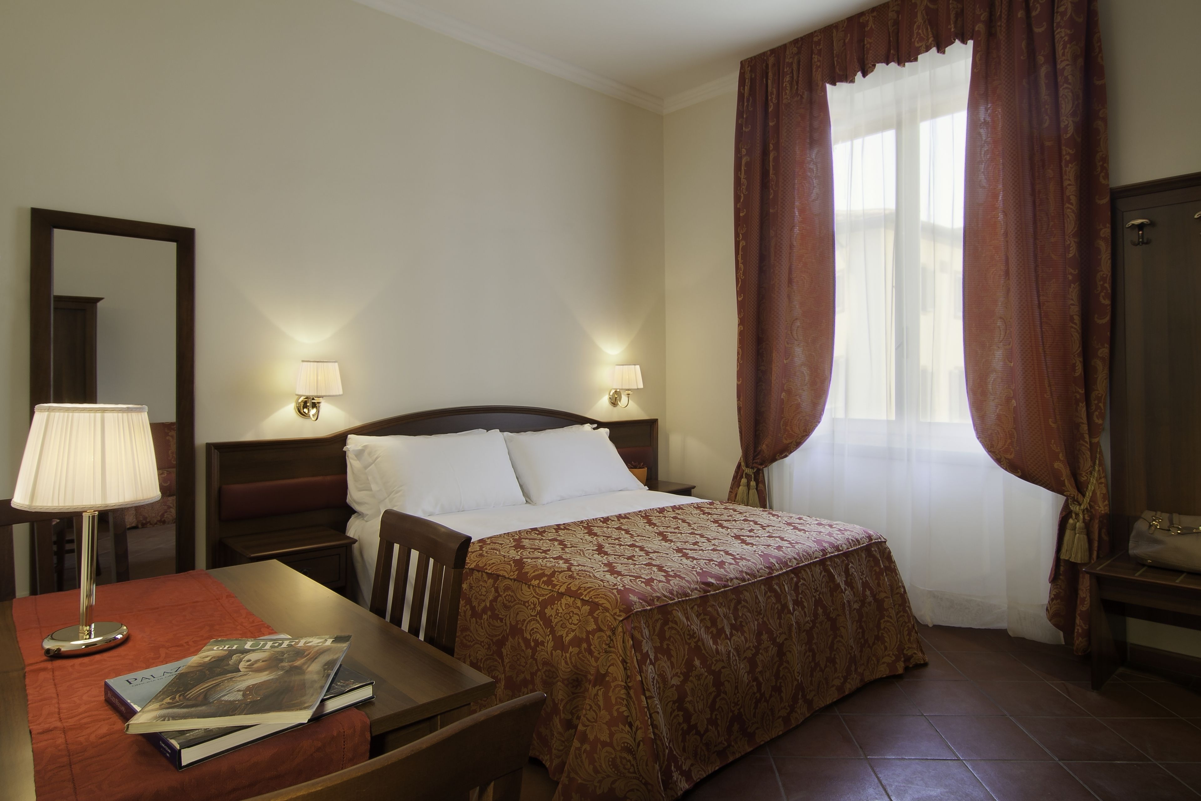 A hotel room with a bed, bedside lamps, a desk with a lamp, a chair, and a window with curtains.
