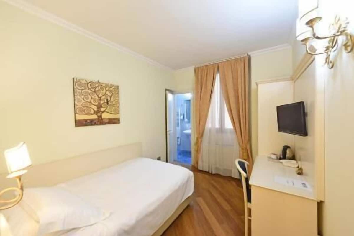 A hotel room with a single bed, a bedside table with a lamp, a television mounted on the wall, and a door leading to a bathroom.