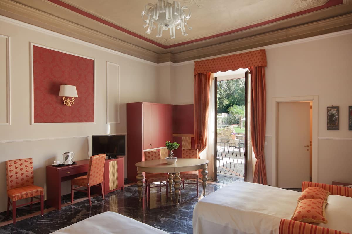 A hotel room with a bed, a desk with a television, a chair, a small table with a plant, and a view of the outdoors.