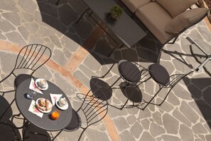 Un área de patio al aire libre con suelo de piedra, sillas de metal, una mesa con una planta y un sofá.