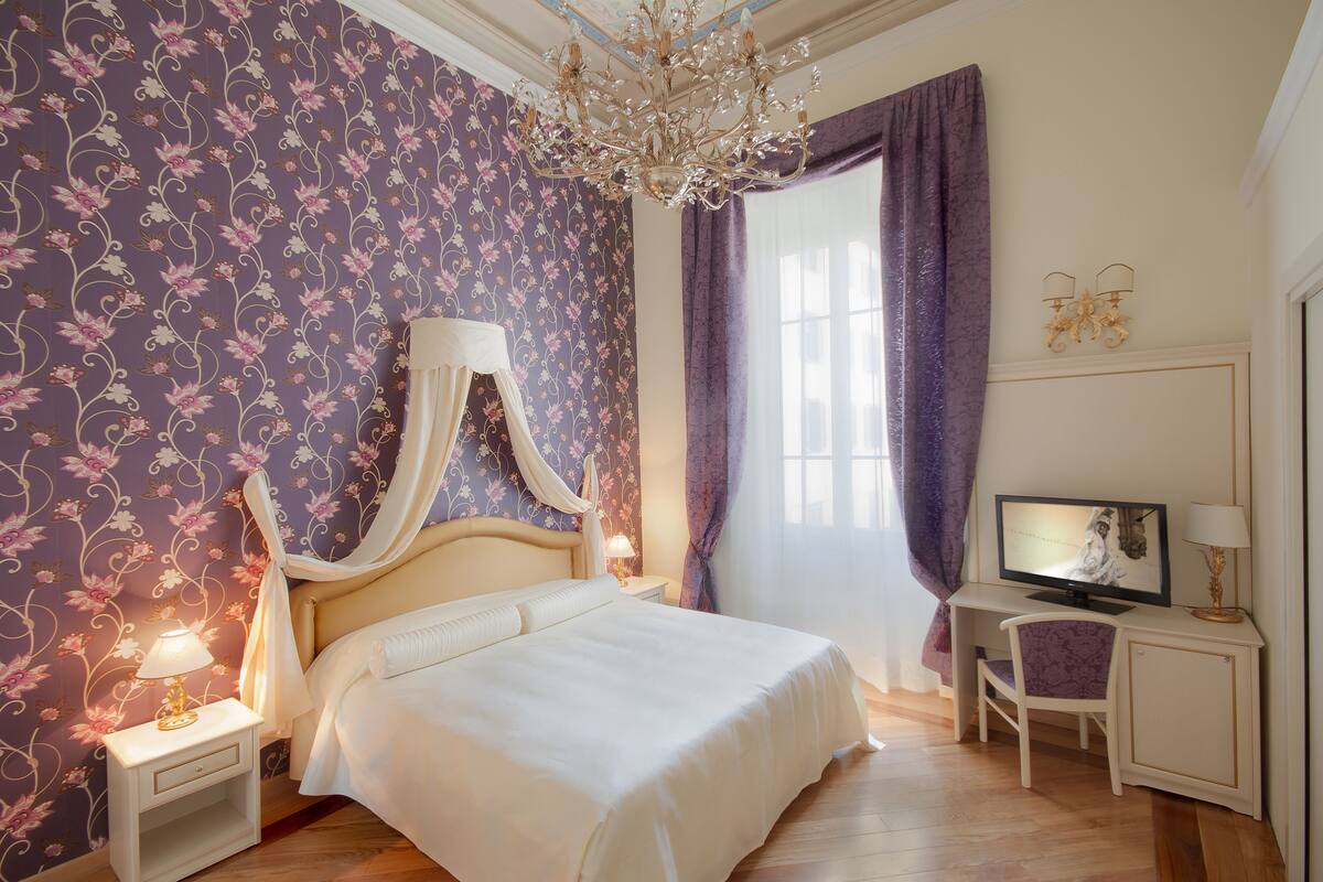 A bedroom with a bed, a nightstand, a desk, a chair, a television, and a chandelier.