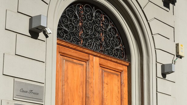 A wooden double door with a circular handle, a black wrought iron decorative arch above it, and a plaque with the text "Casa Fresia" on the wall to the left. - Relais la Corte di Cloris (Florence)