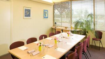 Un salón de conferencias con una mesa larga, sillas, cuadros en la pared y una ventana con persianas.