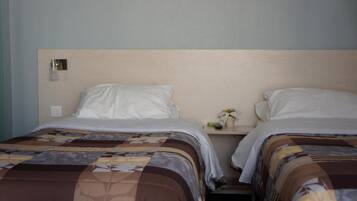 A hotel room with two beds, each with a patterned bedspread, a nightstand with a small plant, and a wall-mounted light fixture.