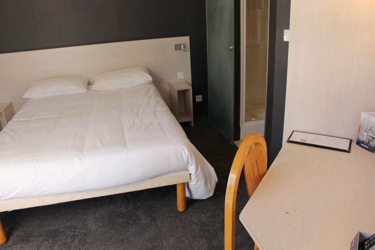 A hotel room with a bed, a small bedside table, a chair, and a desk with a chair.