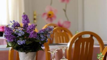 Una mesa de comedor con mantel rosa, una planta en maceta con flores moradas y una jarra de vidrio.