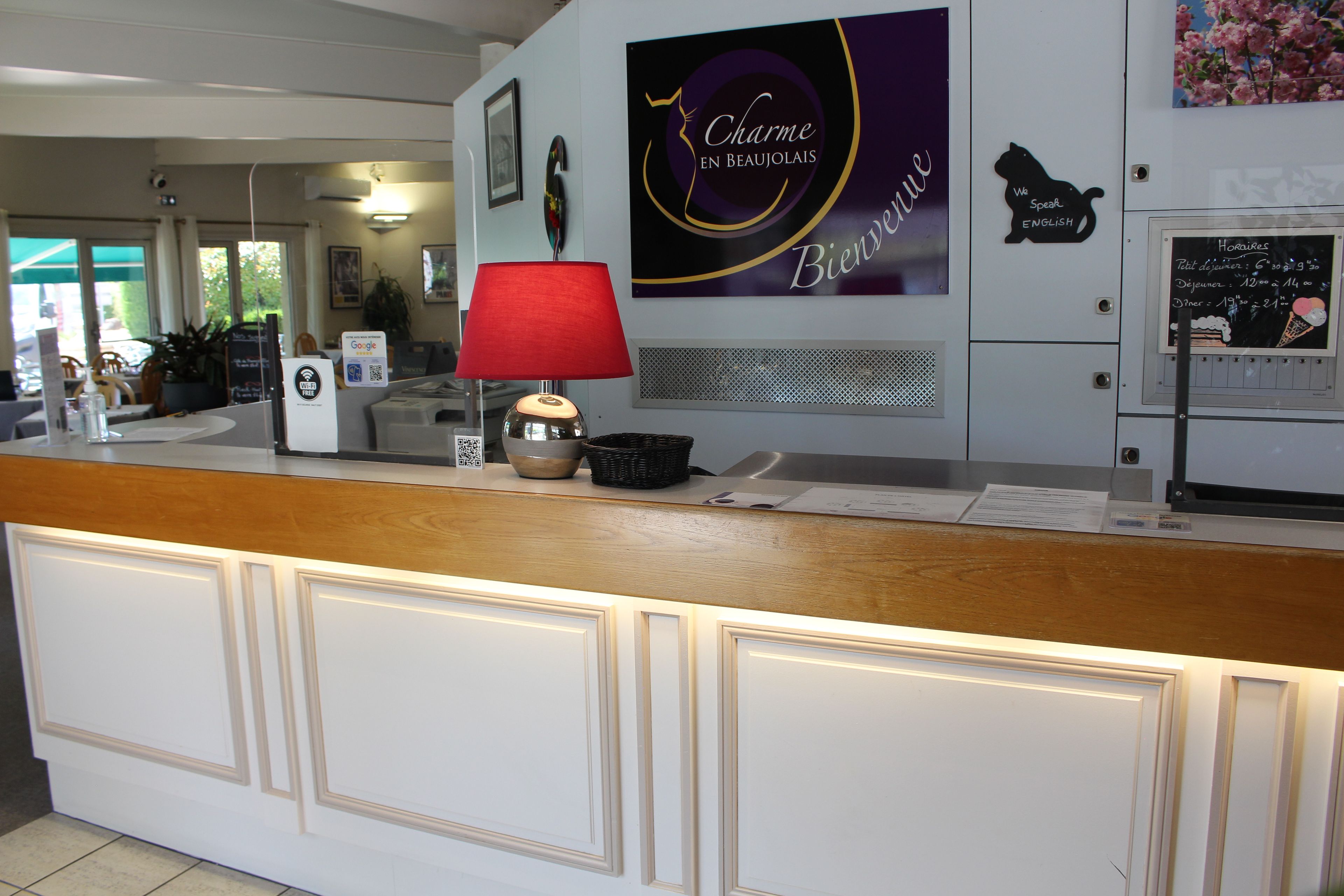 Un comptoir d’accueil d’hôtel avec une lampe rouge, une enseigne indiquant « Charme en Beaujolais » et un panneau affichant le menu.