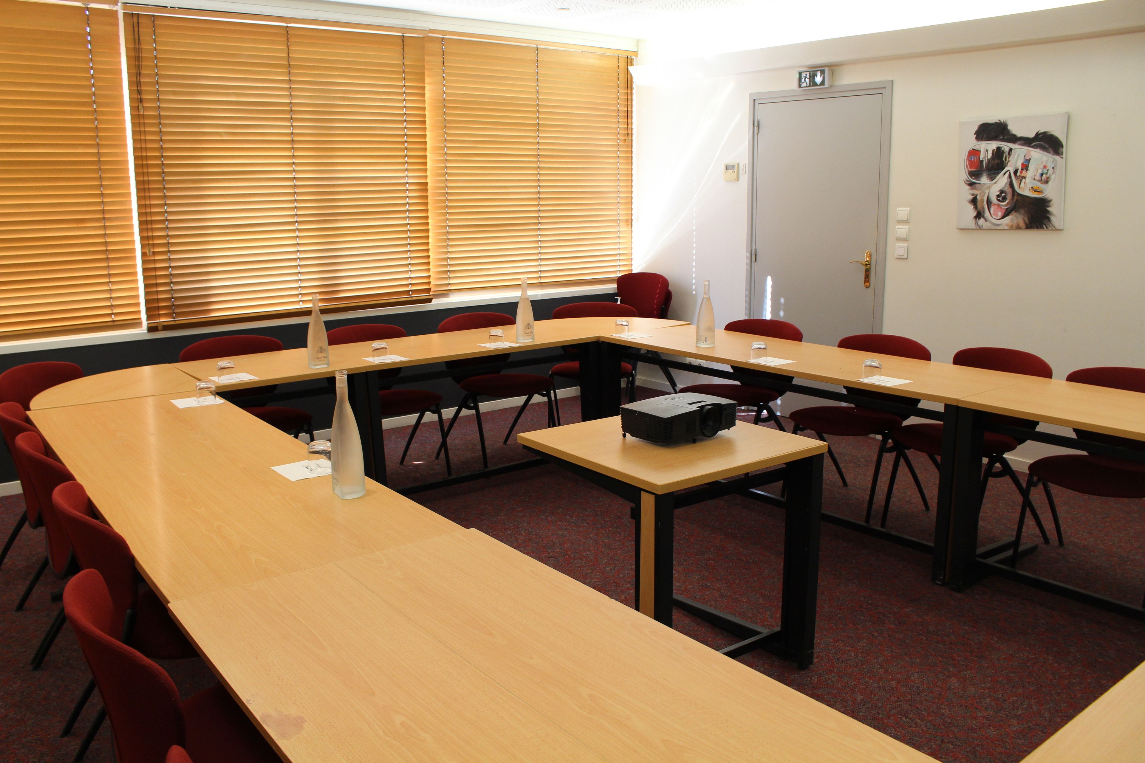 Un salón de conferencias con una larga mesa, sillas rojas, un proyector y una puerta con letrero de salida de emergencia.