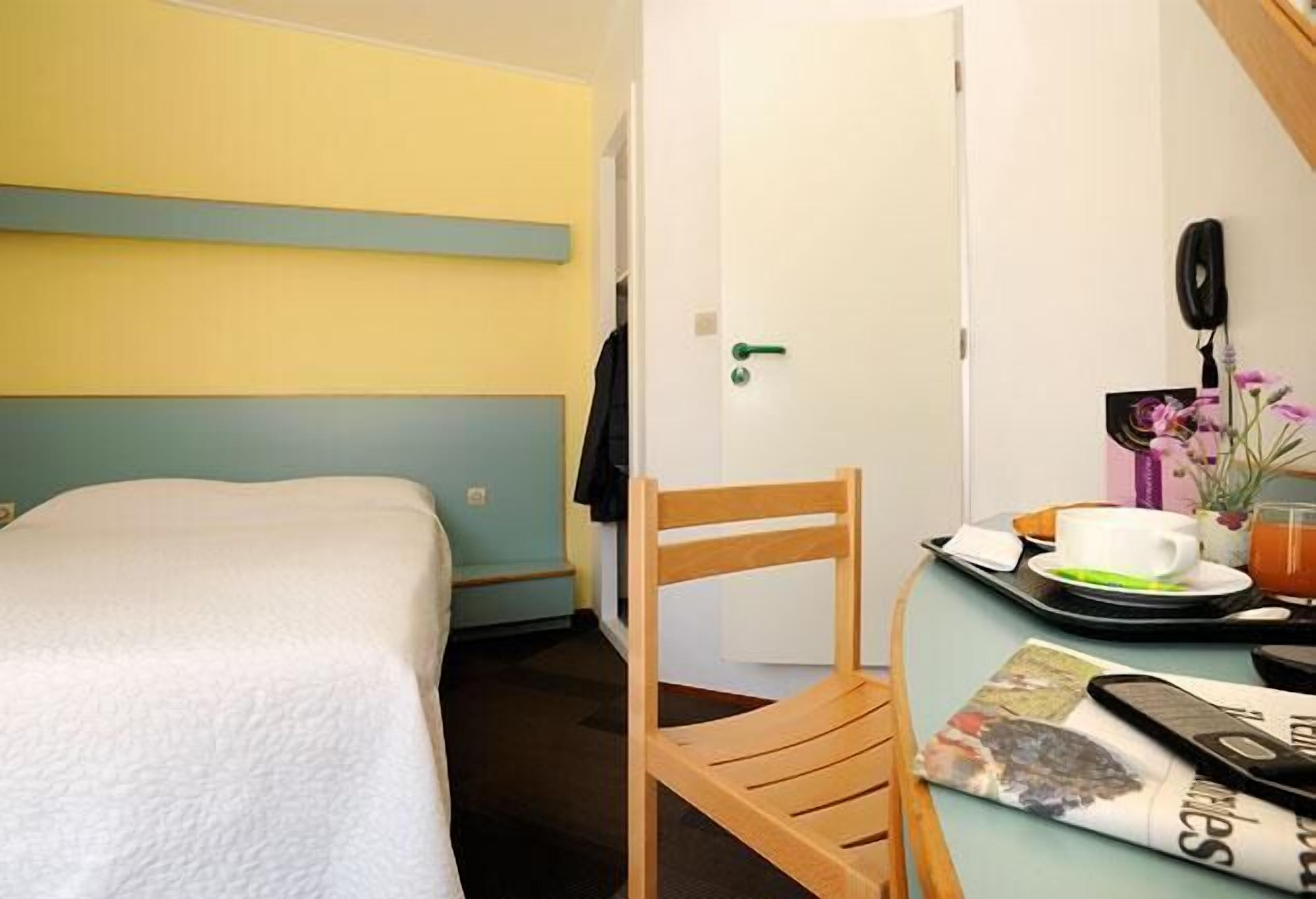 A hotel room with a bed, a wooden chair, a table with a tray, a cup, a phone, and a picture frame.