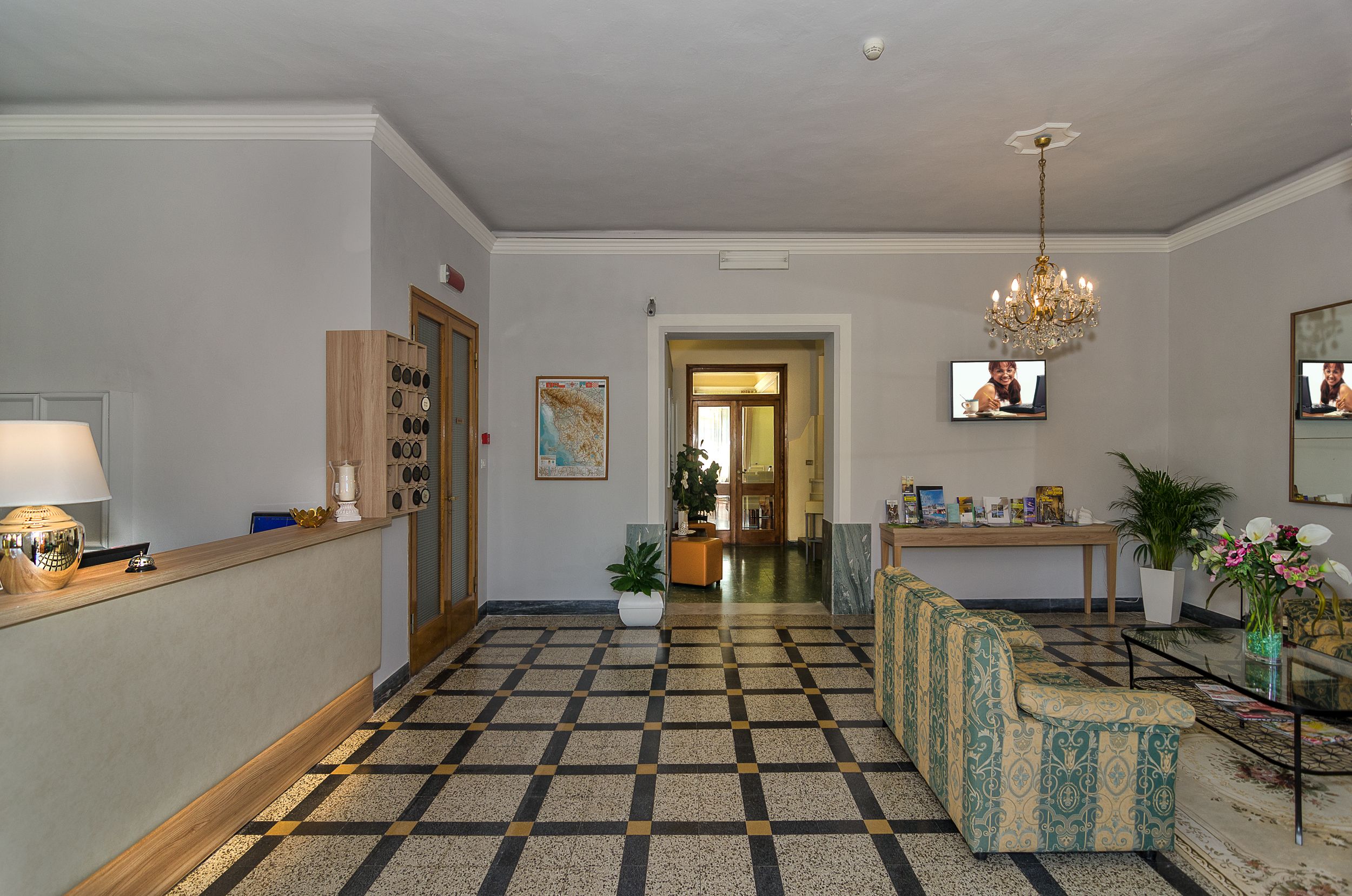 Hall d'hotel con reception, pavimento decorato, angolo salotto e lampadario.