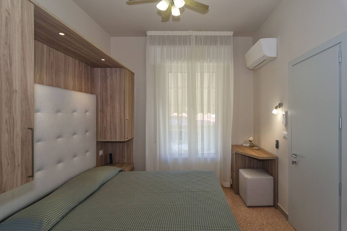 A bedroom with a bed, a bedside table, a small bench, a ceiling fan, and an air conditioning unit.