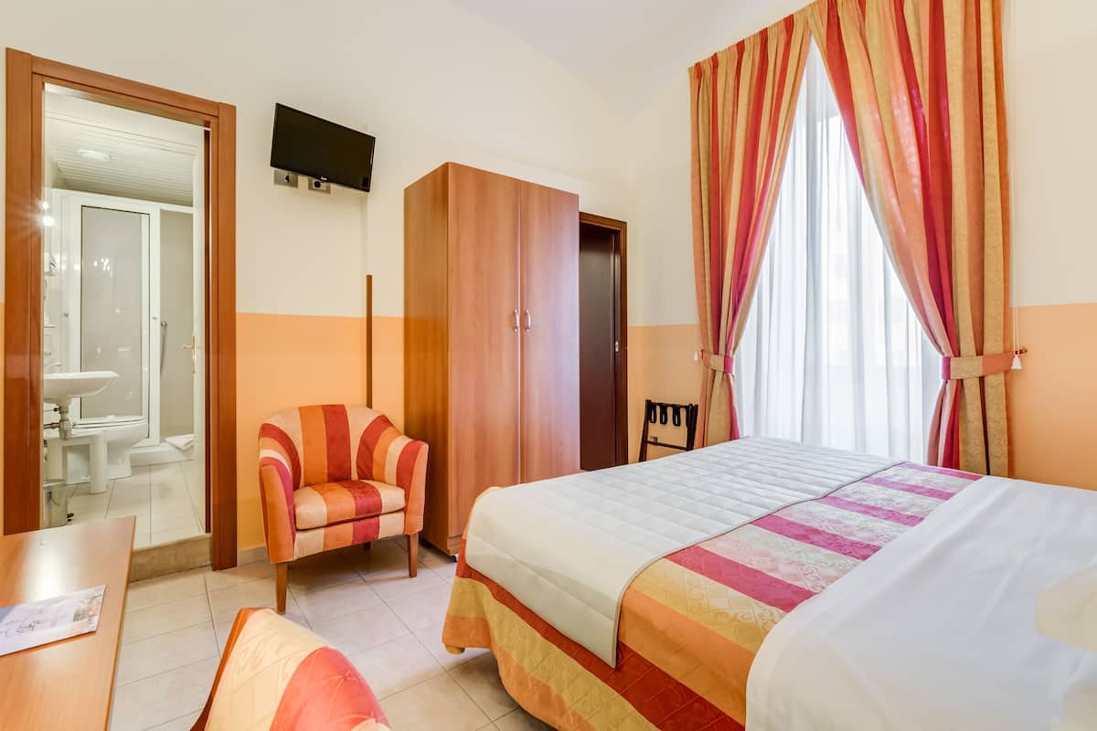 A hotel room with a bed, a chair, a wardrobe, a TV, and a bathroom visible through an open door.