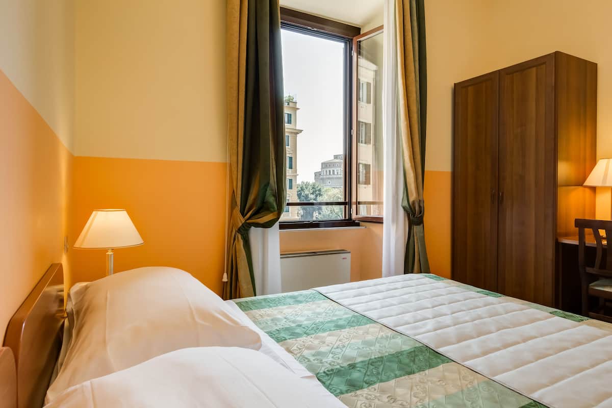 A hotel room with a bed, a window with curtains, a radiator, and a wooden wardrobe.