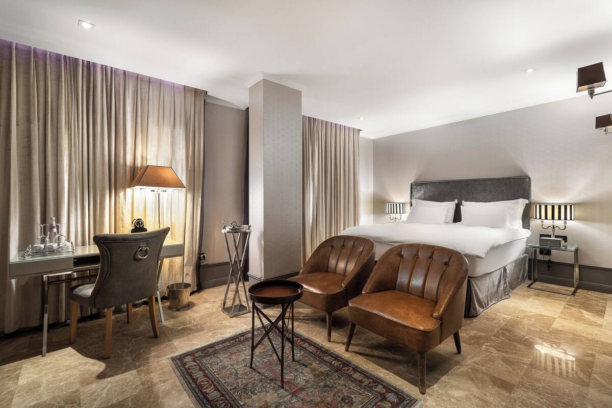 A hotel room with a bed, two leather chairs, a desk with a lamp, and a small table with a lamp.