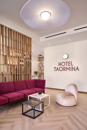 A modern hotel lobby with a red sofa, a round chair, a coffee table, and a wall-mounted sign reading 'HOTEL TAORMINA'. - Hotel Taormina (Bardolino)