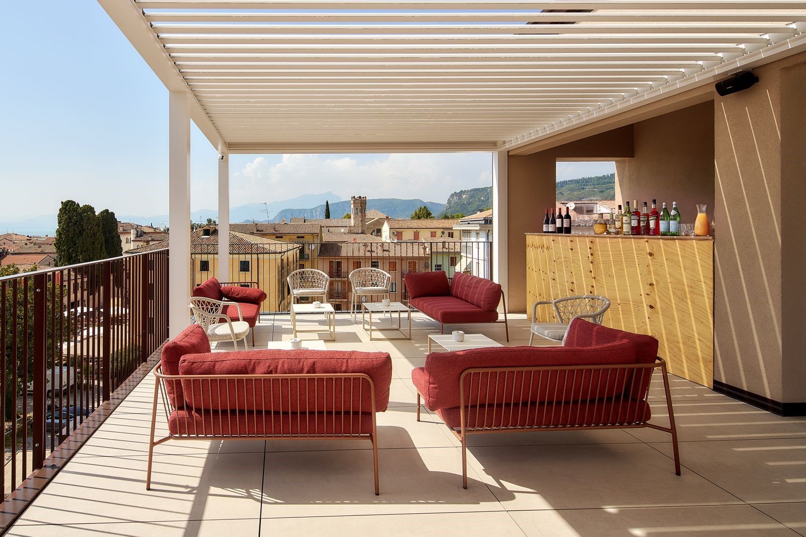 Una terraza con sofás rojos y sillas blancas, una barra de madera con botellas, y vistas a edificios y montañas.