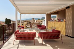 A terrace with red sofas and white chairs, a wooden bar with bottles, and a view of buildings and mountains. - Hotel Taormina (Bardolino)