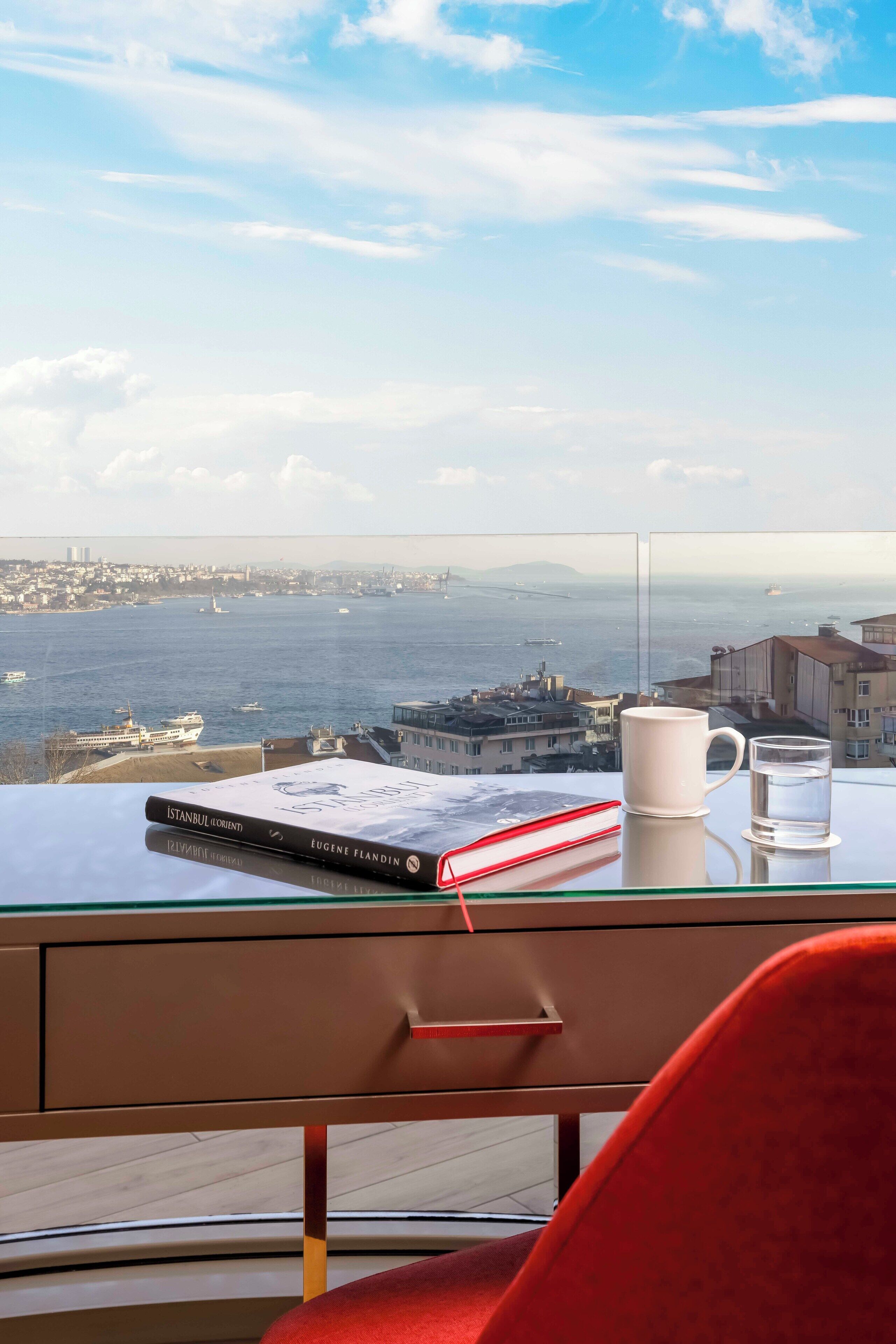 A view from a room with a glass table displaying a book and a mug, overlooking a body of water with boats and buildings.