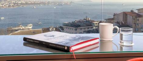 A view from a room with a glass table displaying a book and a mug, overlooking a body of water with boats and buildings.