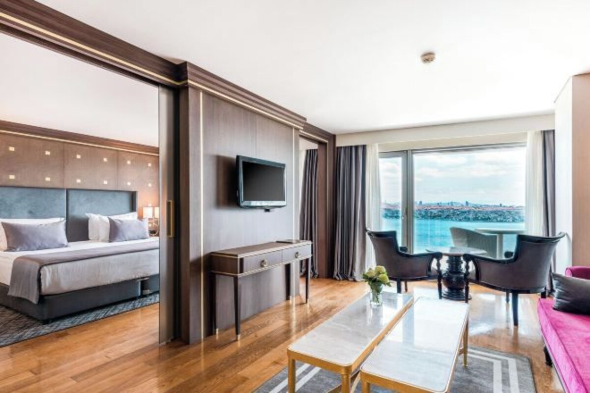 A modern hotel room with a bed, a TV, a dining table, and a view of the city.