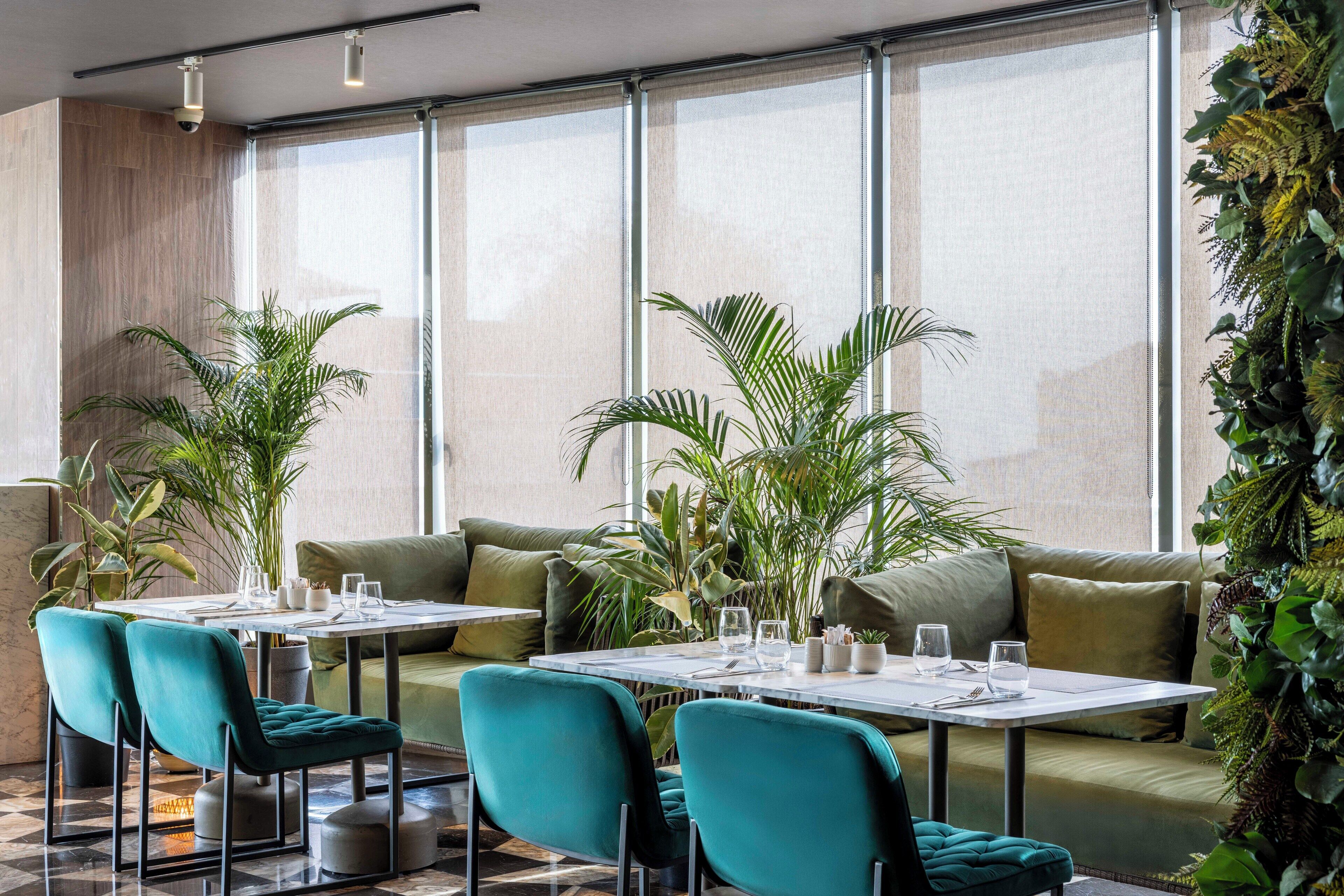 A modern dining area with green upholstered chairs, a white table, and a green sofa. There are potted plants and a large window with blinds.