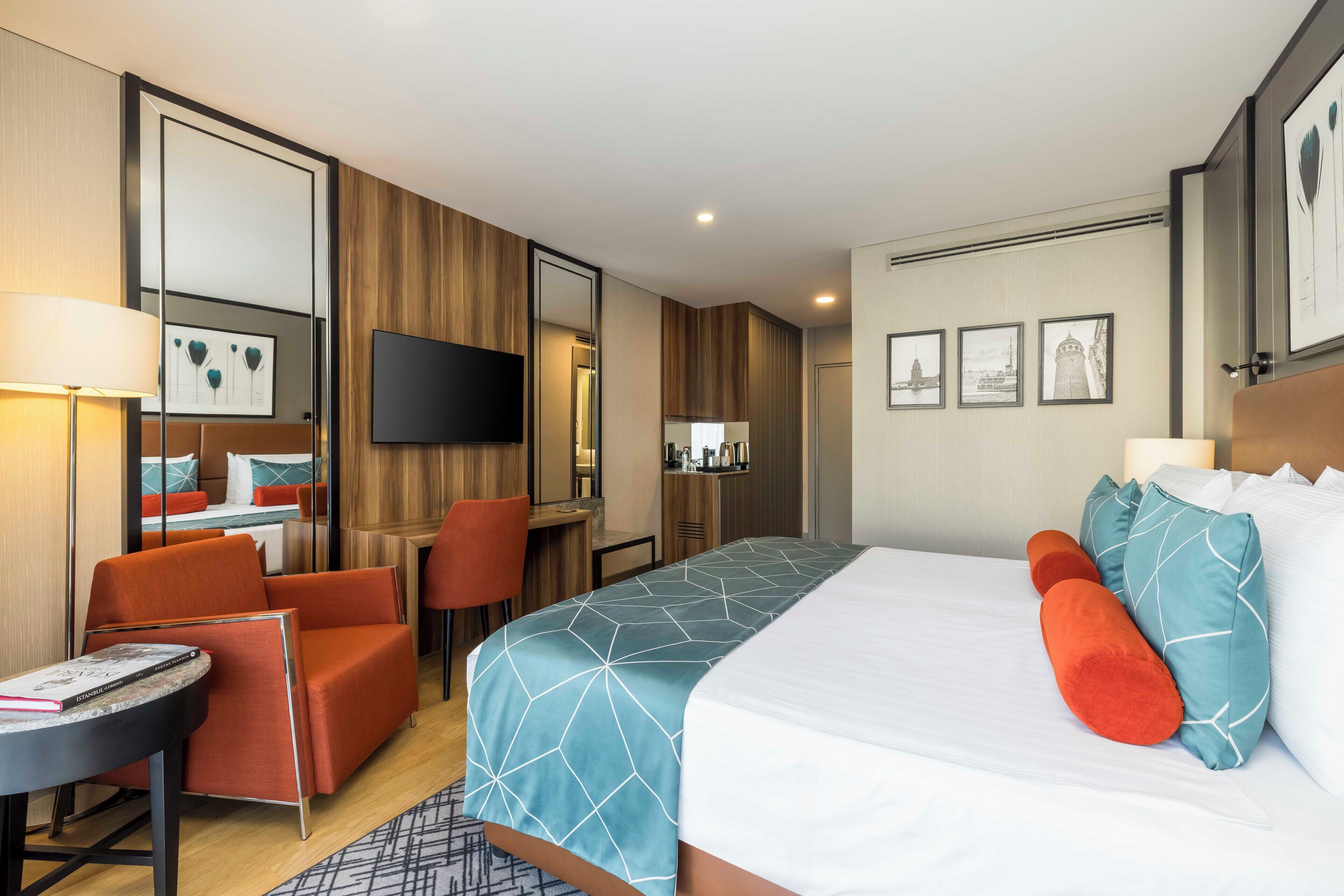 A hotel room with a bed, orange armchair, desk, and a TV on the wall.