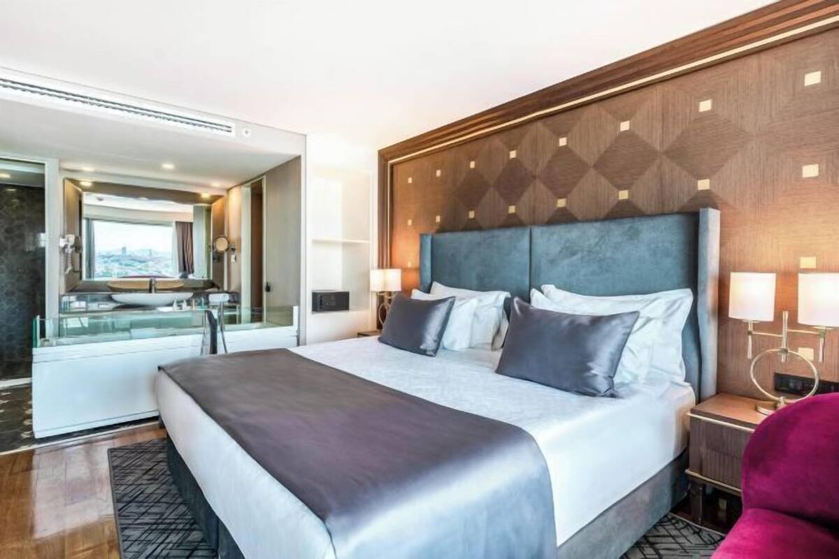 Quarto de hotel moderno com uma cama grande, banheiro com paredes de vidro e vista para a cidade.
