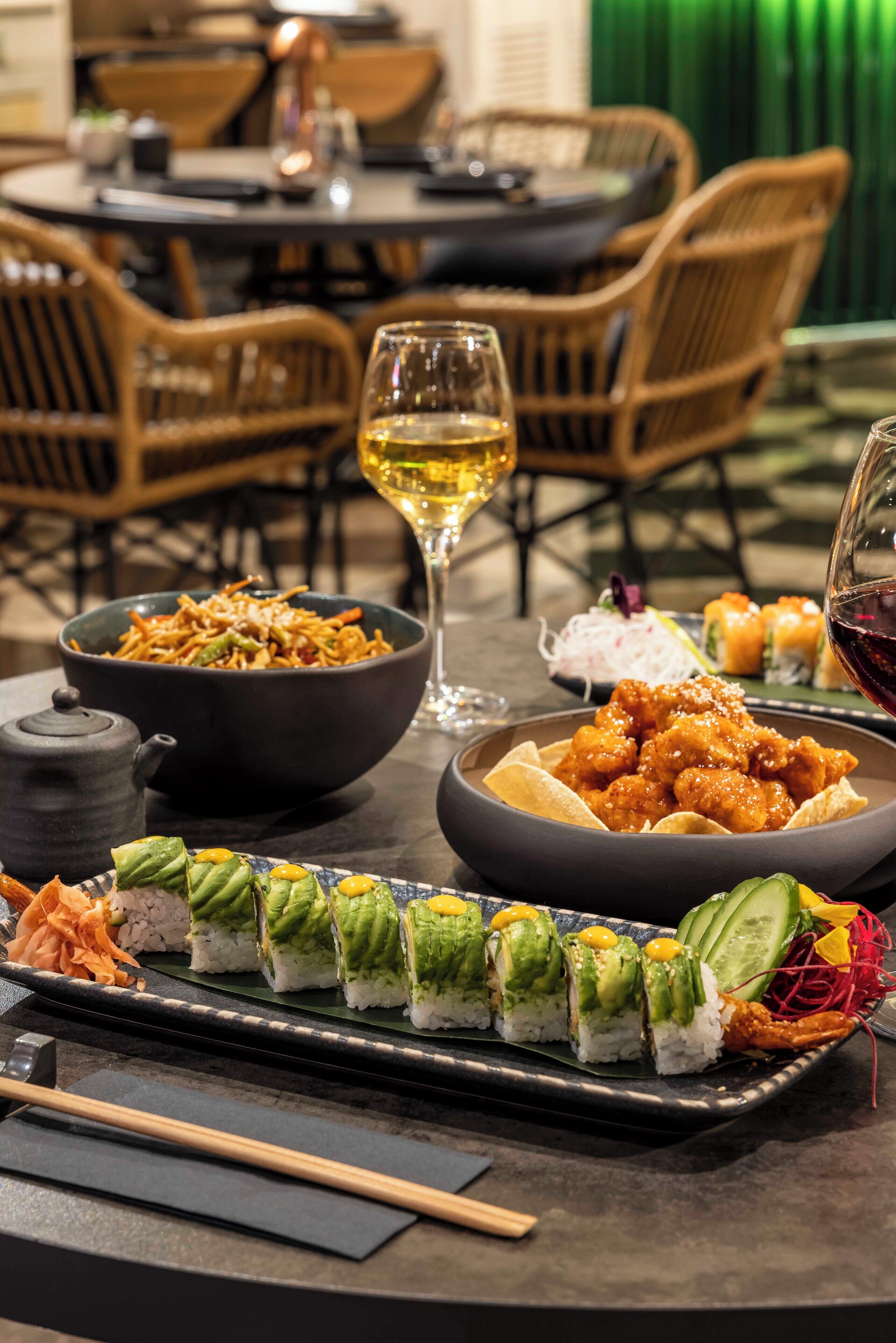 Une table à manger dressée avec une variété de plats, notamment des sushis, un bol de nouilles et un verre de vin.