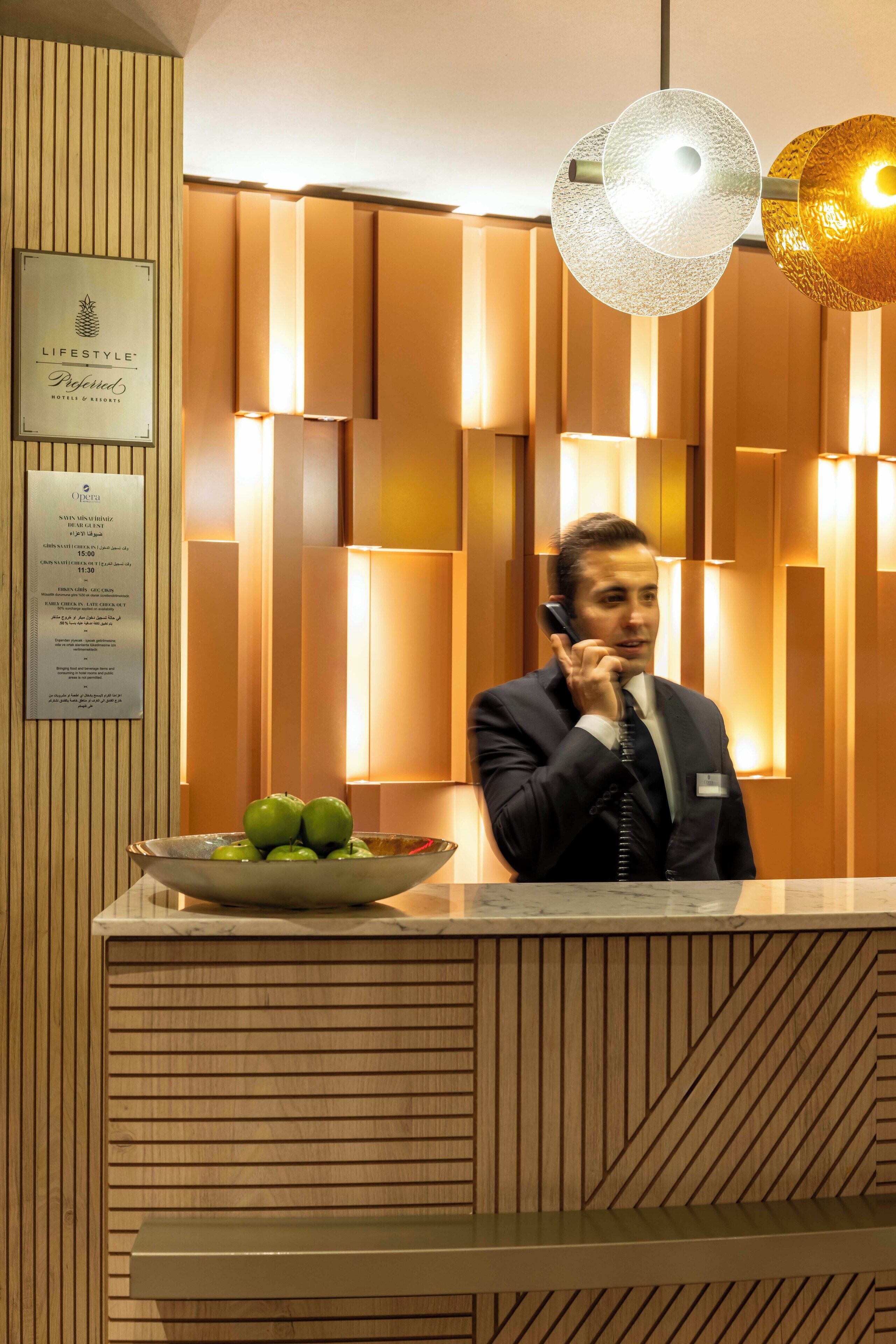 A man in a suit at a reception desk with a bowl of green apples, wooden paneling, and a sign for 'Lifestyle Preferred Hotels & Resorts'.