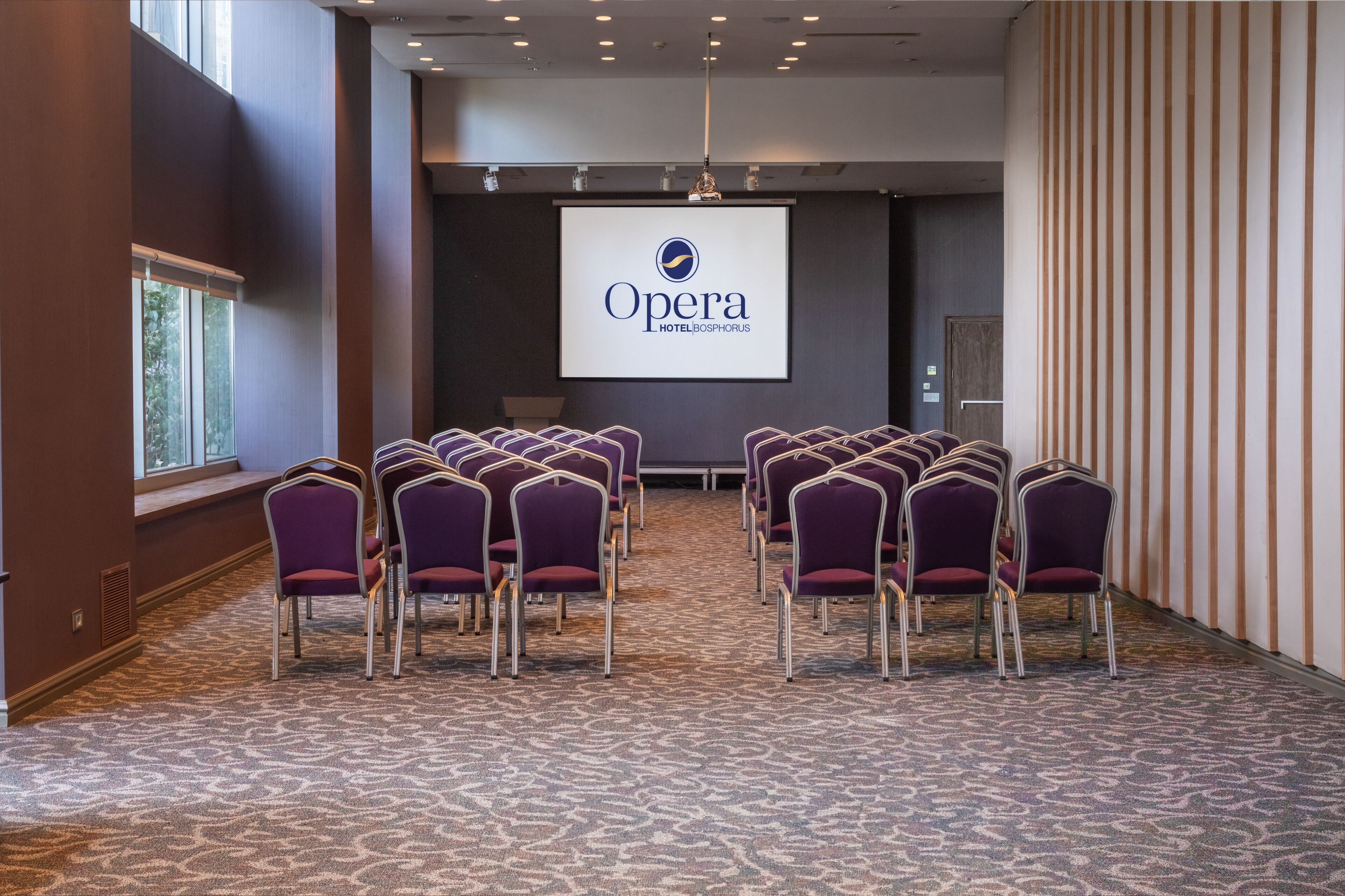 A conference room with rows of purple chairs, a projector screen displaying the Opera Hotel logo, and a patterned carpet.