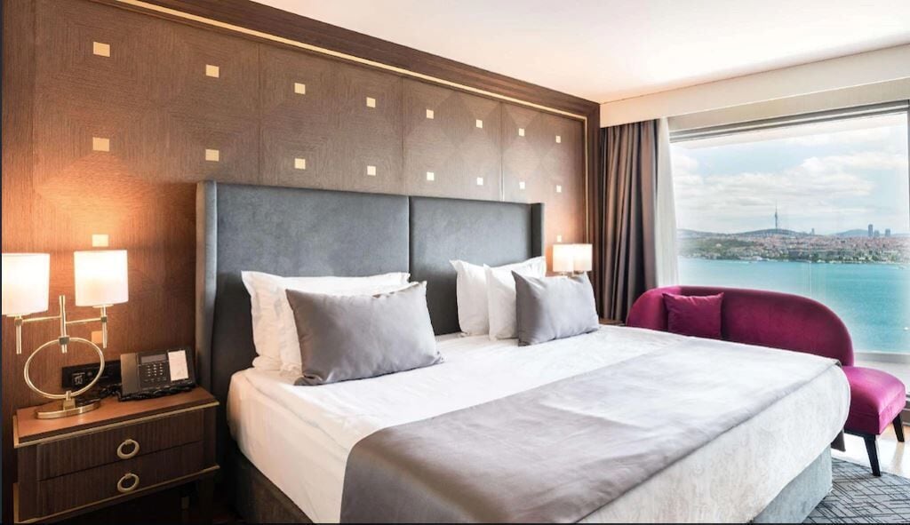A hotel room with a large bed, two bedside tables with lamps, a view of a cityscape through a large window, and a city skyline visible outside the window.