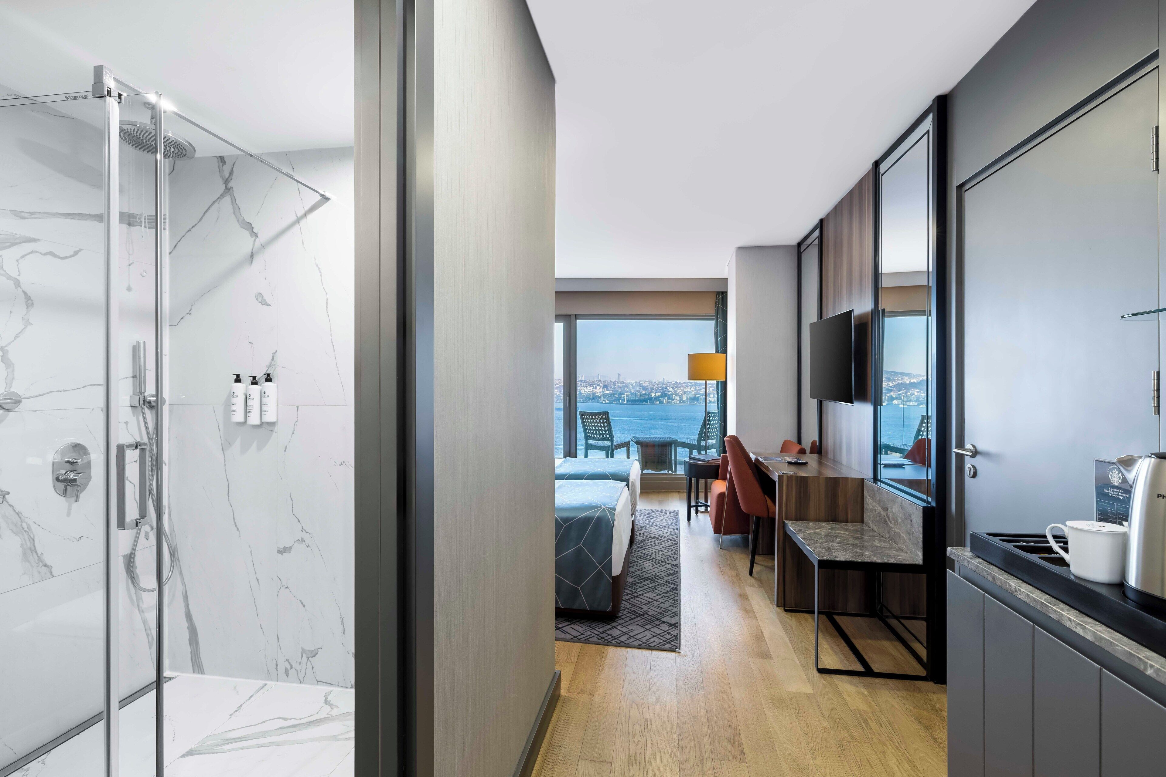 A modern hotel room with a glass-enclosed shower, a bed with a geometric-patterned bedspread, a dining area with a table and chairs, and a large window offering a city view.