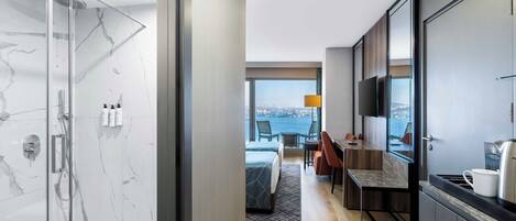 A modern hotel room with a glass-enclosed shower, a bed with a geometric-patterned bedspread, a dining area with a table and chairs, and a large window offering a city view.