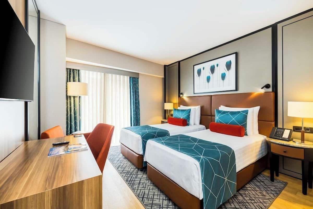 Quarto de hotel com duas camas, uma escrivaninha, uma televisão e uma janela com cortinas.