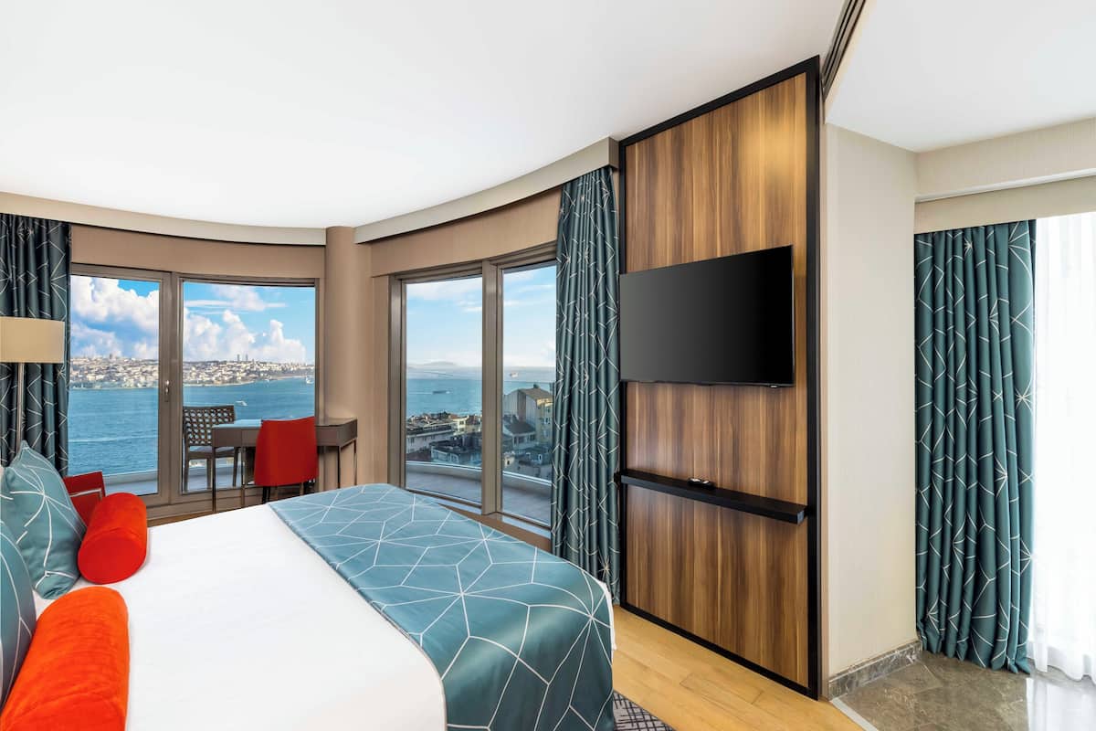 Quarto de hotel com cama grande, televisão na parede, varanda com vista para o mar e área de refeições com cadeiras vermelhas.
