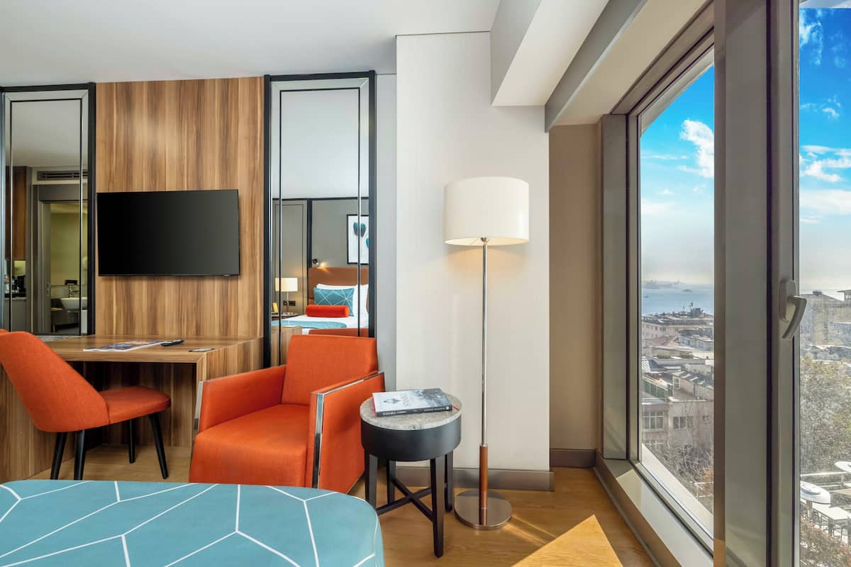 Quarto de hotel com uma janela grande, uma televisão de tela plana, uma poltrona laranja, uma mesa redonda pequena com um livro e uma cama com colcha de padrão geométrico.