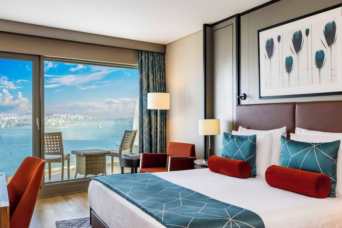Quarto de hotel com uma cama grande, duas cadeiras, uma mesinha e vista para o mar.
