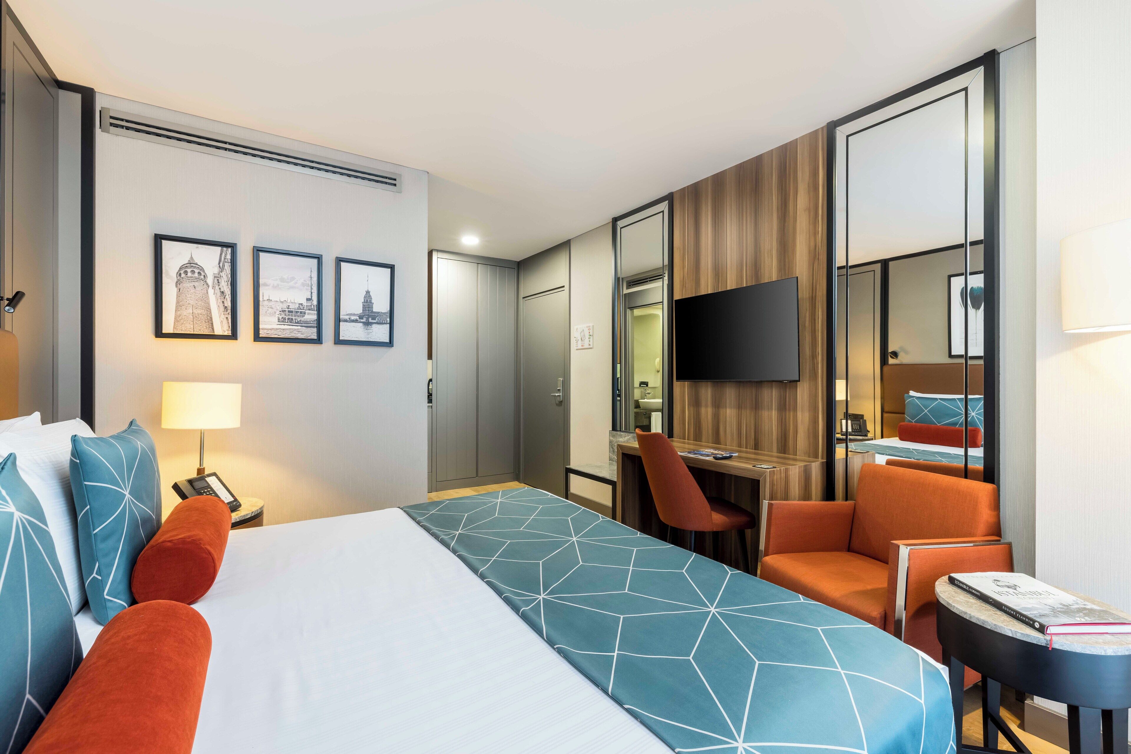 A hotel room with a large bed, a desk, a chair, a TV, and a small table with a book.