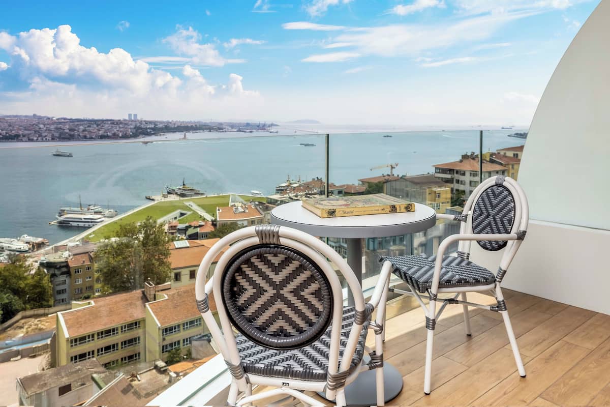 Varanda com duas cadeiras de vime e uma mesa pequena, com vista para um porto e edifícios.