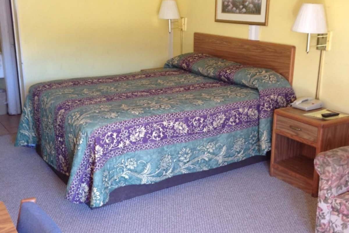 A single bed with a patterned bedspread, a nightstand with a lamp, a chair, and a framed picture on the wall.