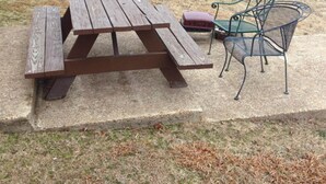 Une table de pique-nique avec des bancs et une chaise, située dans un espace herbeux, avec un abri et des arbres en arrière-plan.