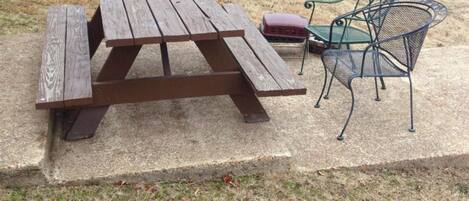 Una mesa de picnic con bancos y una silla en un área verde, con un cobertizo y árboles al fondo.