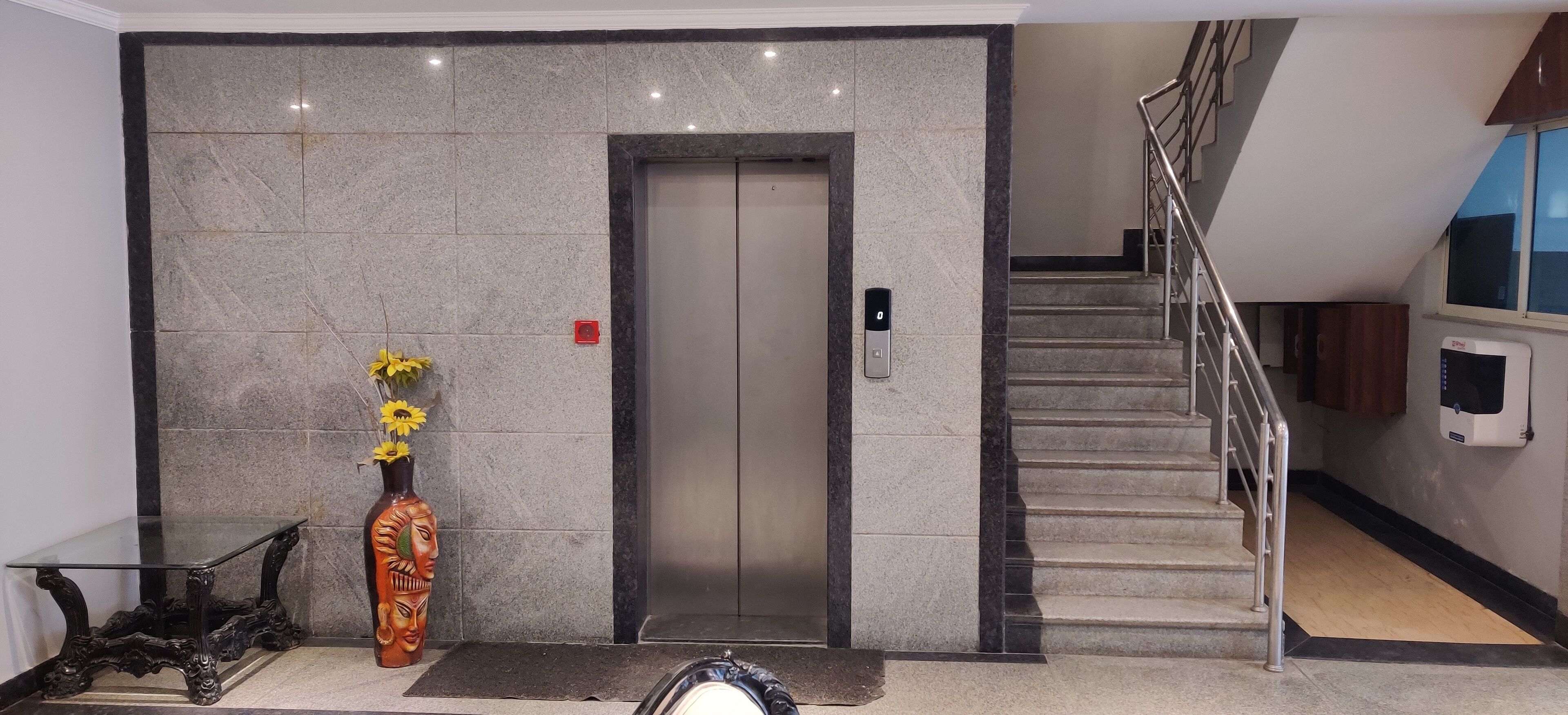 Un vestíbul modern amb una paret de marbre, una taula de vidre, un vas amb flors, un ascensor i una escala.