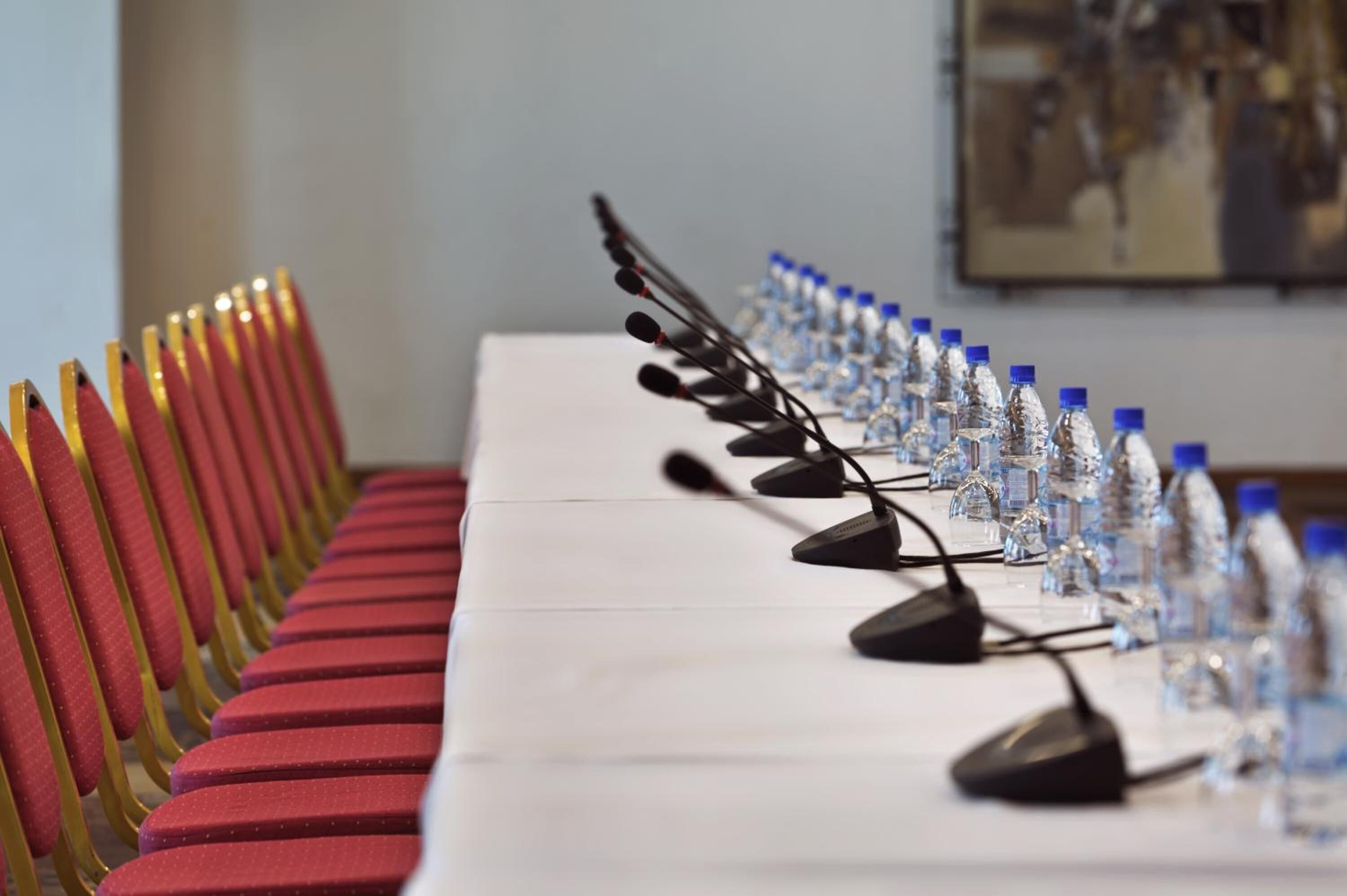 Una mesa de conferencias con micrófonos y botellas de agua, sillas rojas con detalles amarillos, y un cuadro en la pared.