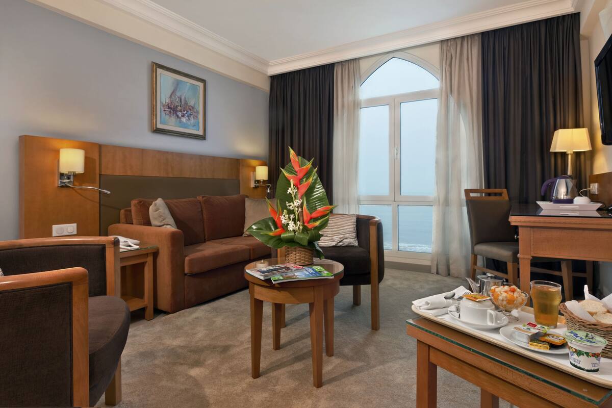 A hotel room with a brown sofa, a wooden coffee table with a magazine, a tray with breakfast items, and a large window with curtains.