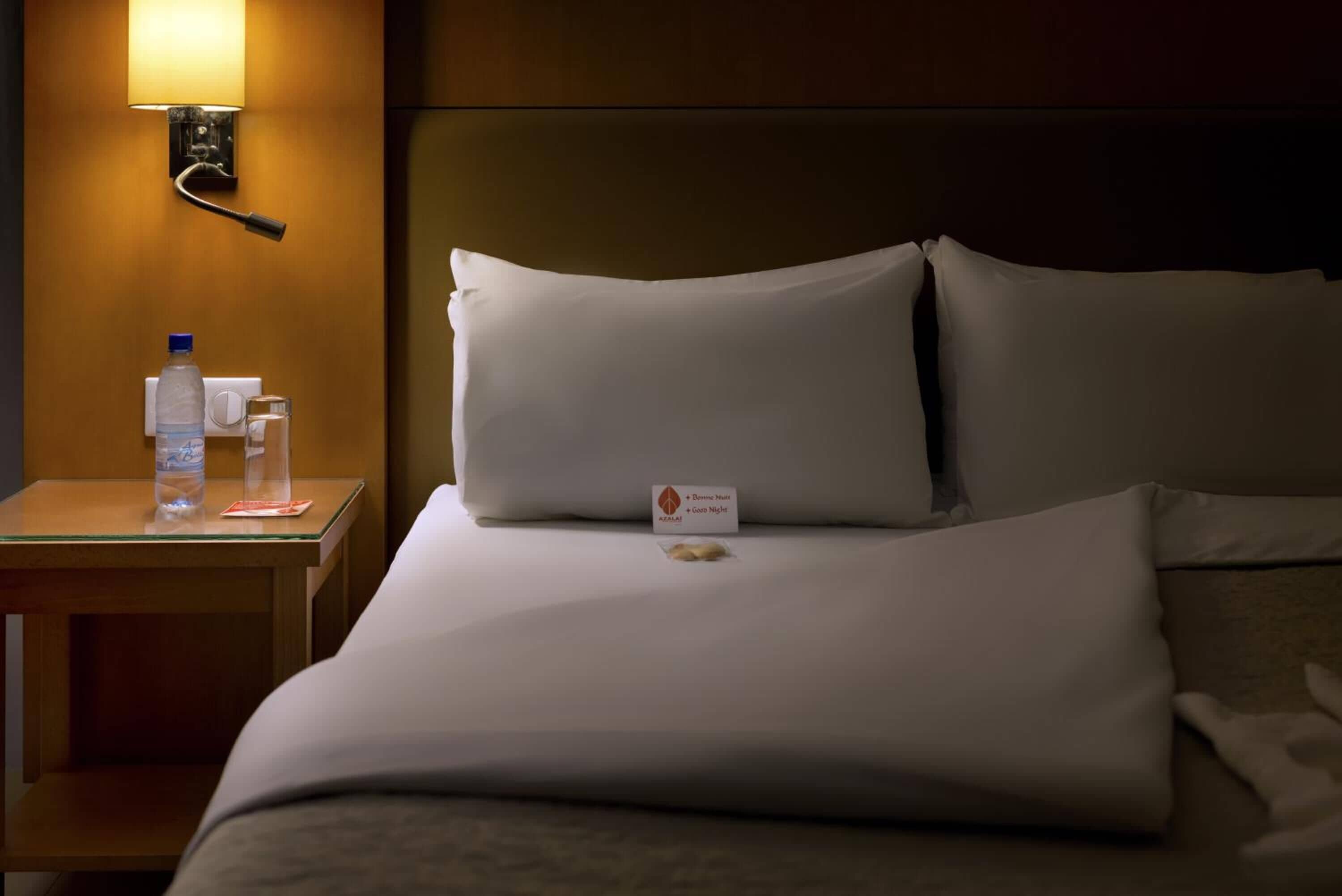A hotel room with a neatly made bed, a bedside table with a water bottle and glass, and a wall-mounted lamp.