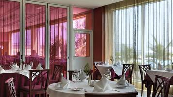 A restaurant with round tables and chairs, large windows with curtains, and a view of palm trees.
